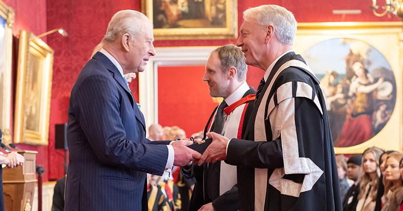The King in a blue suit shakes hands with the Vice-Chancellor Professor Chris Day who is wearing a full ceremonial black and gold robe.